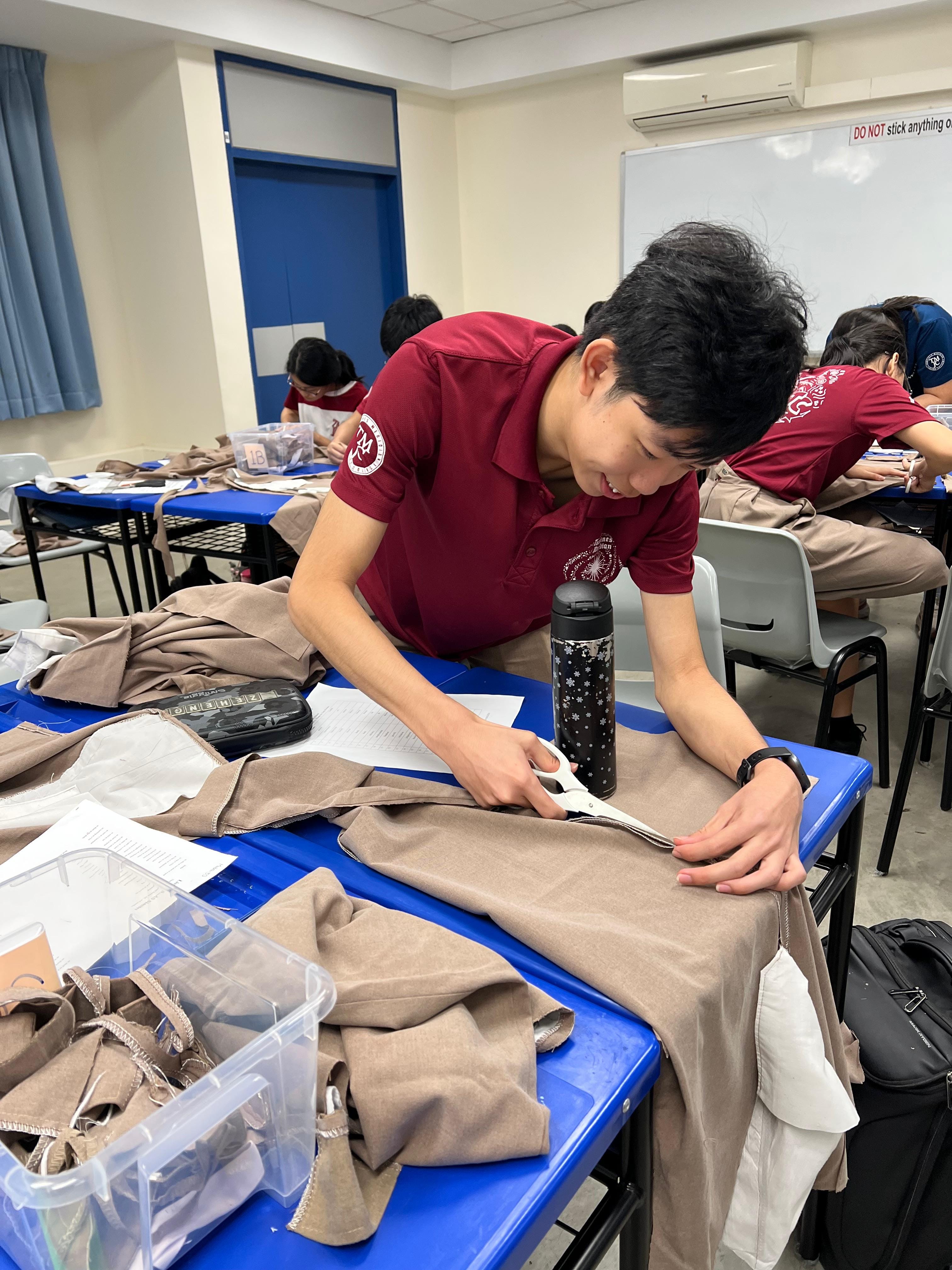 A student in a red polo shirt carefully cuts fabric with scissors at a blue desk. Other students work in the background in the same classroom.